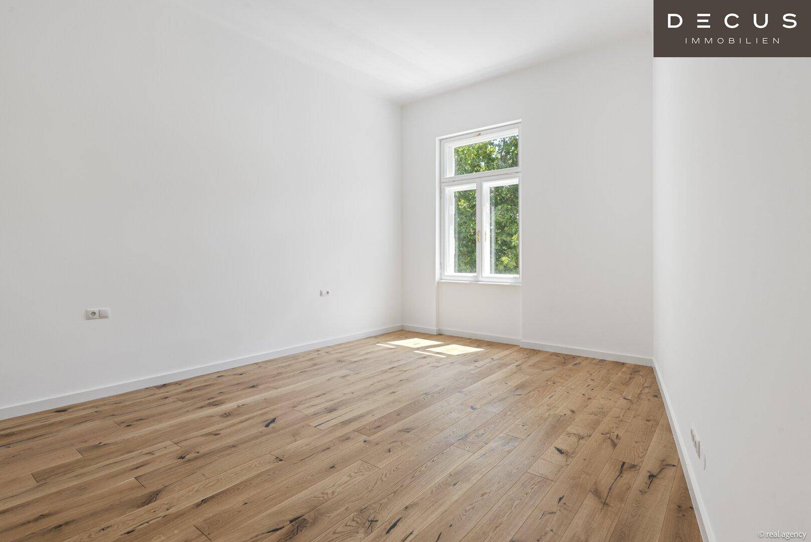 Ruhiges Zimmer mit hellem Holzparkett, weißen Wänden und einem Fenster mit Grünblick.