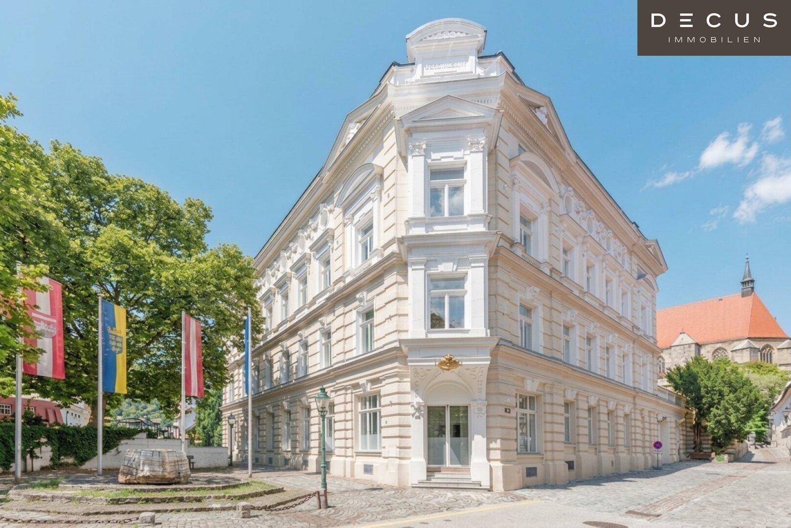 Historisches Eckgebäude mit prächtiger Fassade, umgeben von Bäumen und einer Kirche im Hintergrund.
