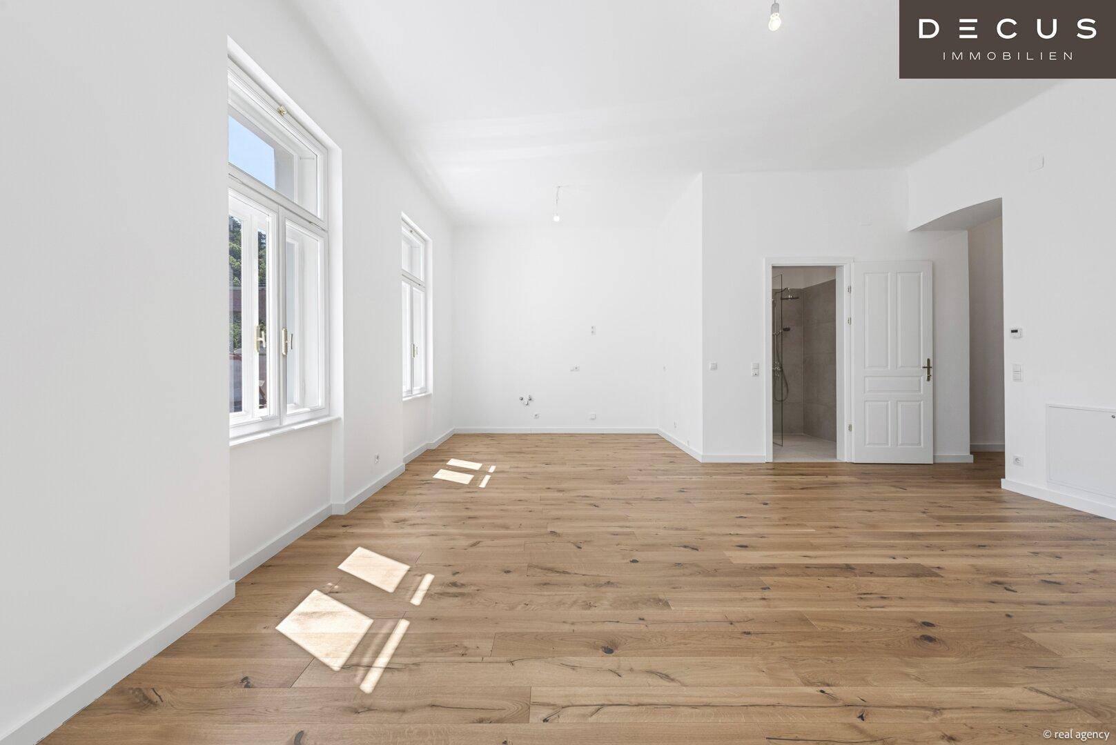 Großer, heller Raum mit Holzboden und hohen Fenstern, die viel natürliches Licht hereinlassen.