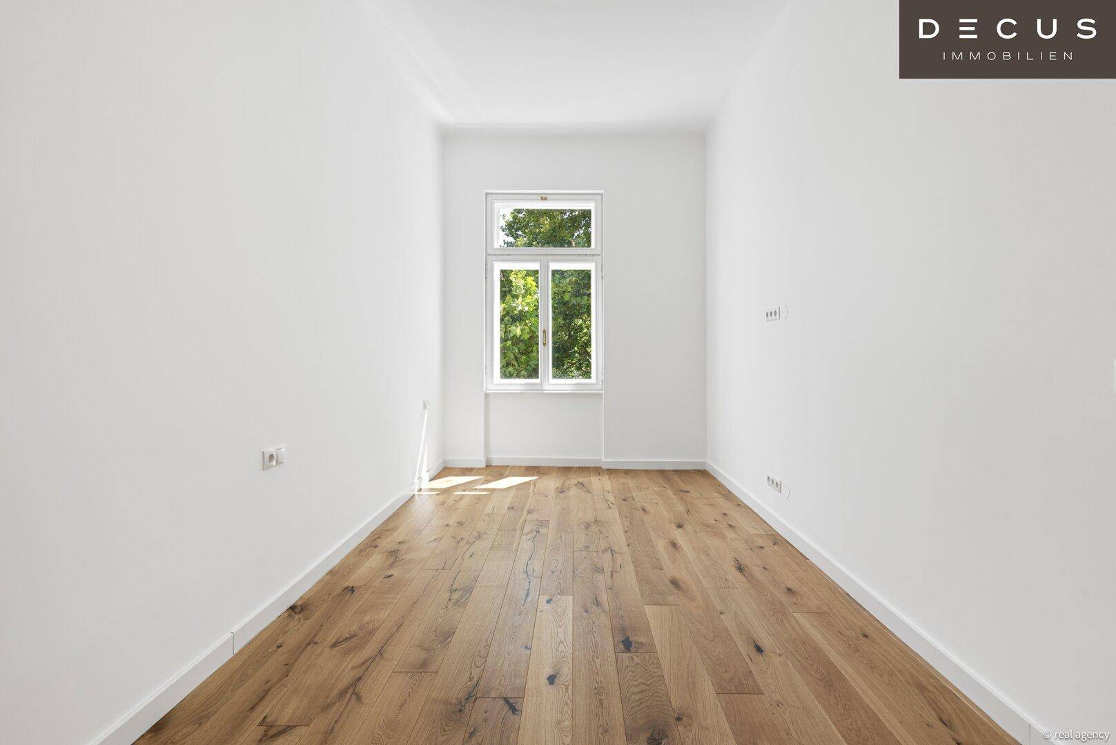Gemütlicher, heller Raum mit Holzdielenboden und einem Fenster mit Blick ins Grüne.