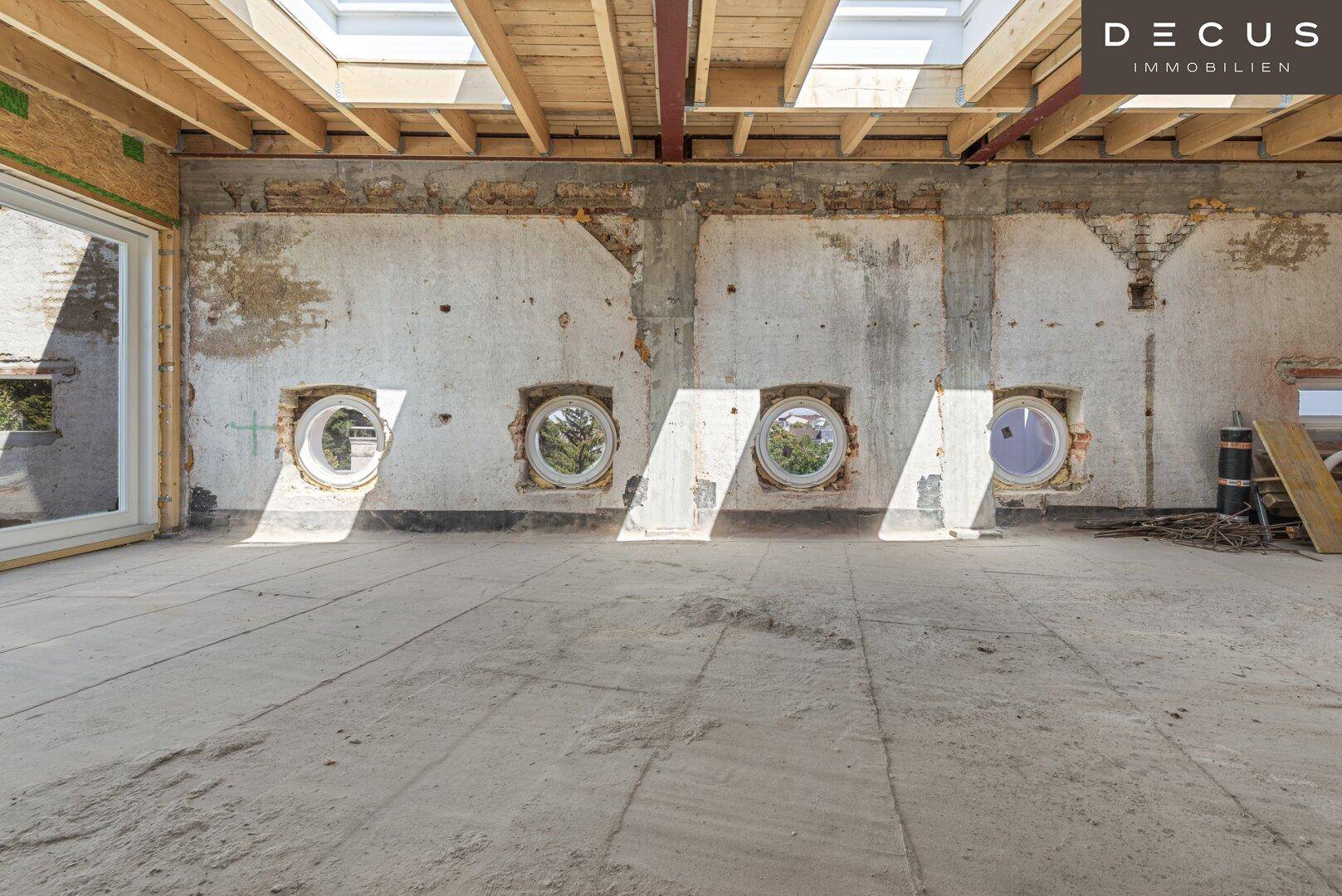 Großer, lichtdurchfluteter Innenraum im Rohbau mit runden Fenstern und Dachfenstern.