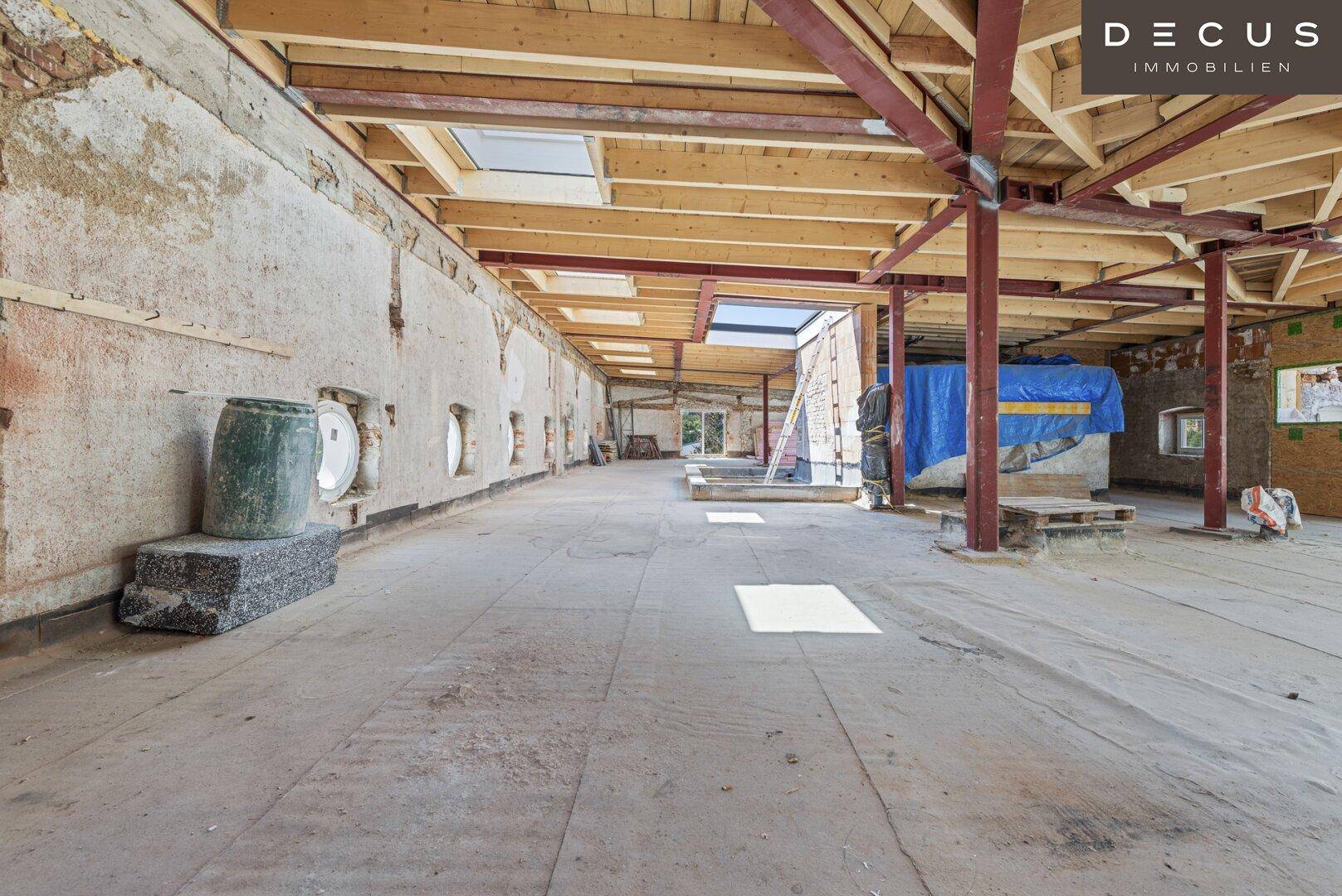 Langer Innenraum im Rohbau mit runden Fenstern, Holzbalken und Betonboden.