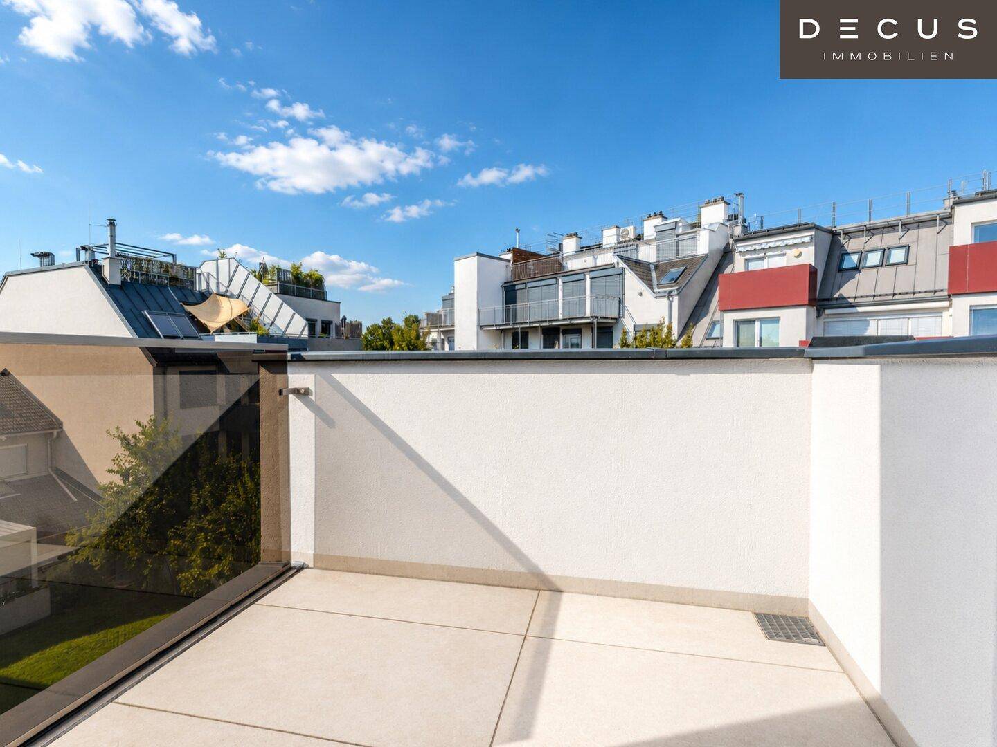 Geräumige Dachterrasse mit Fliesenboden und Glasgeländer, umgeben von anderen Gebäuden.