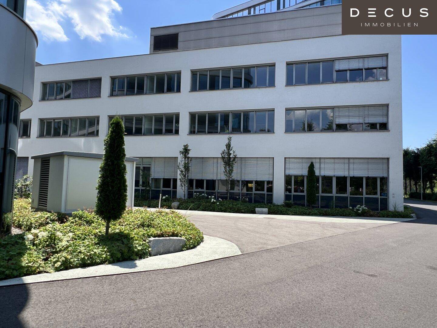 Moderne Bürogebäude-Fassade mit großen Fenstern und gepflegter Außenanlage unter blauem Himmel.
