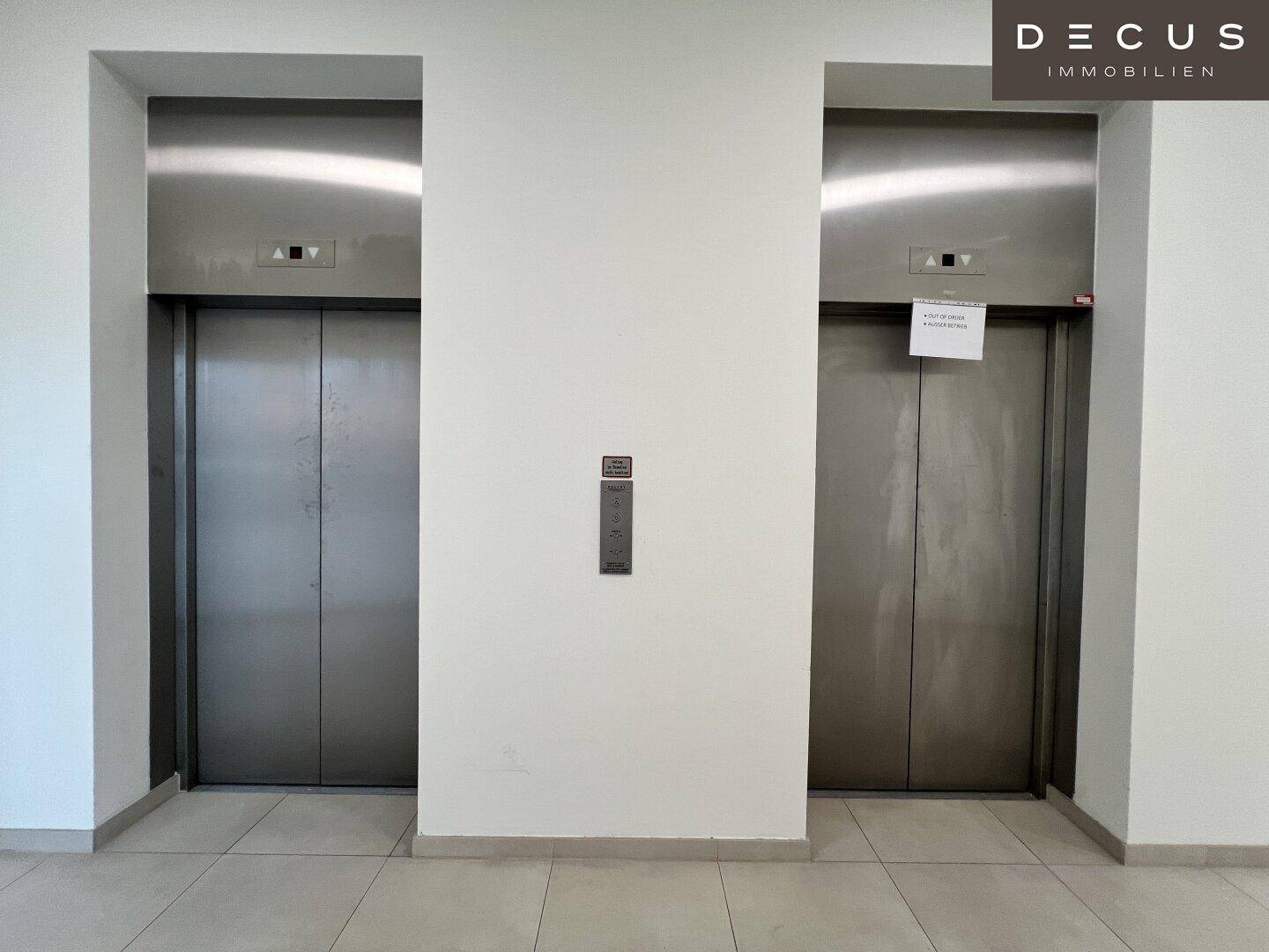 Zwei moderne Aufzüge mit Edelstahltüren in einem hellen, gut gepflegten Flur mit Fliesenboden.