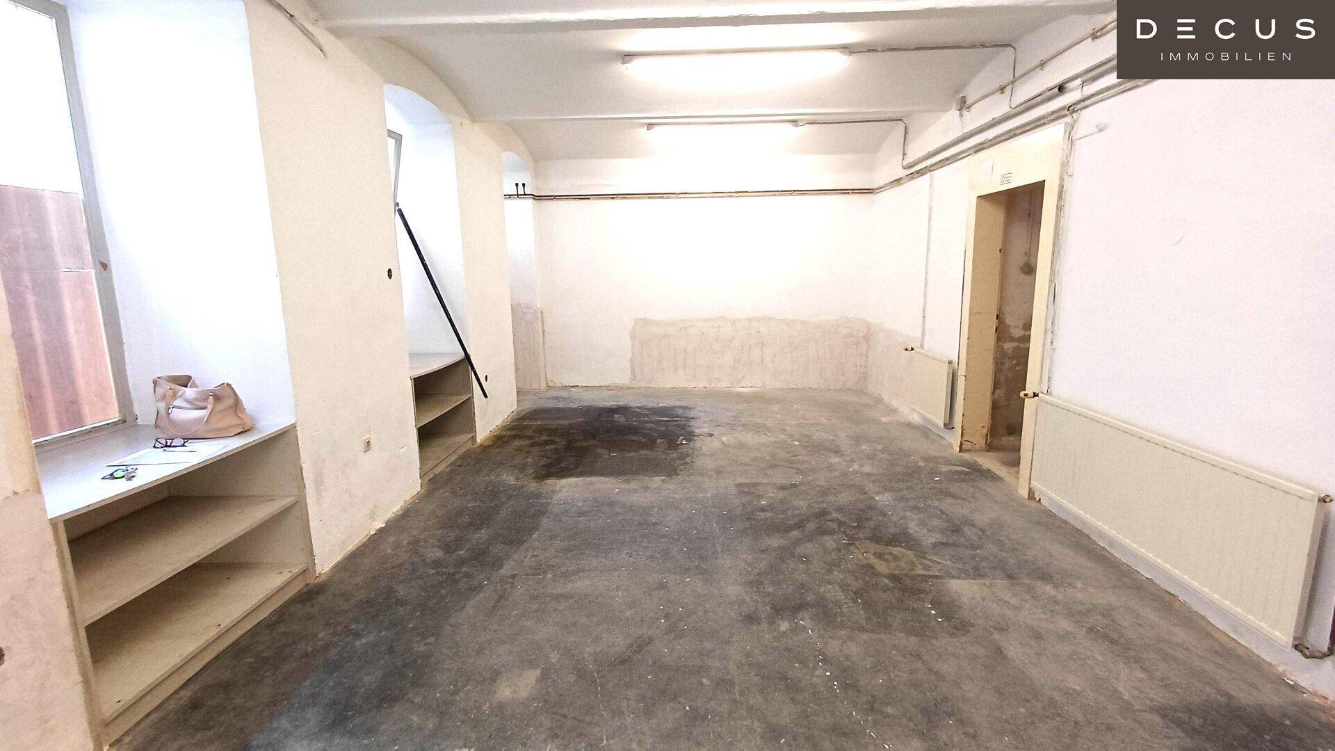 Lagerraum oder Kellerbereich mit Betonboden, sichtbaren Rohren und eingebauten Regalen unter den Fenstern.