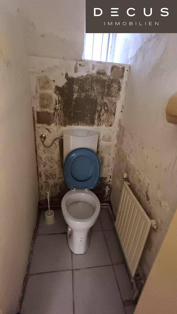 Kleine Toilette mit WC, Heizkörper und Fliesenboden, die Wände sind sanierungsbedürftig.