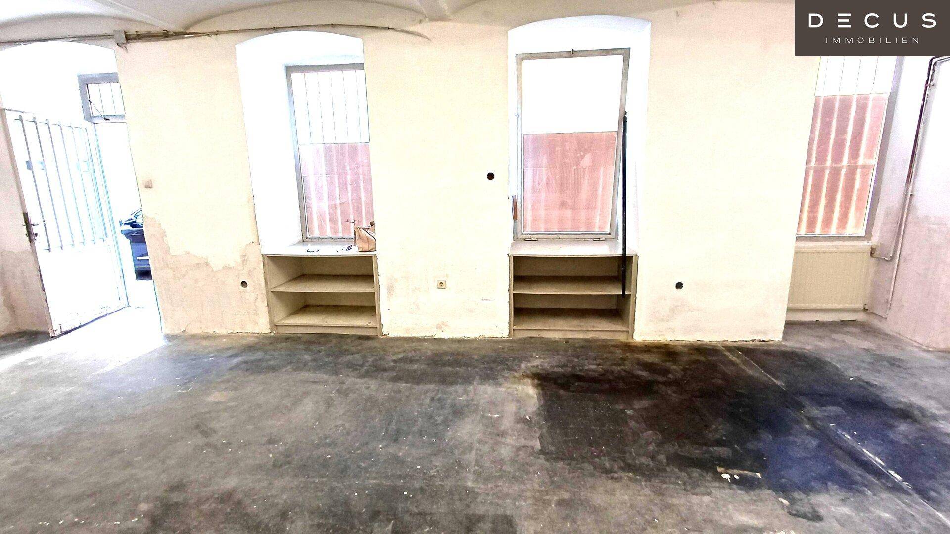 Lagerraum mit Betonboden, eingebauten Regalen unter den Fenstern und sanierungsbedürftigen Wänden.