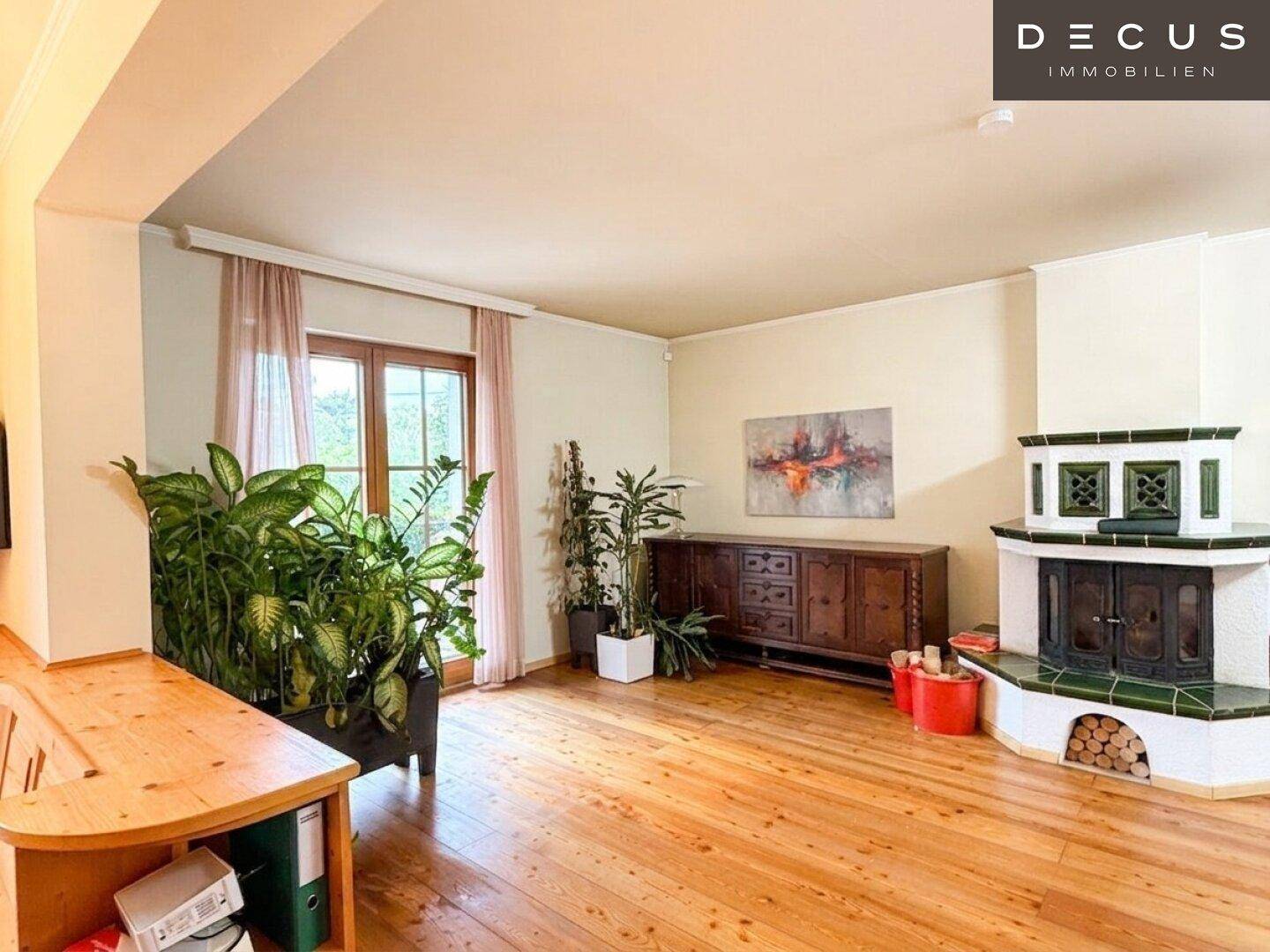 Geräumiges Wohnzimmer mit hellem Holzfußboden, Kachelofen und Blick ins Grüne.