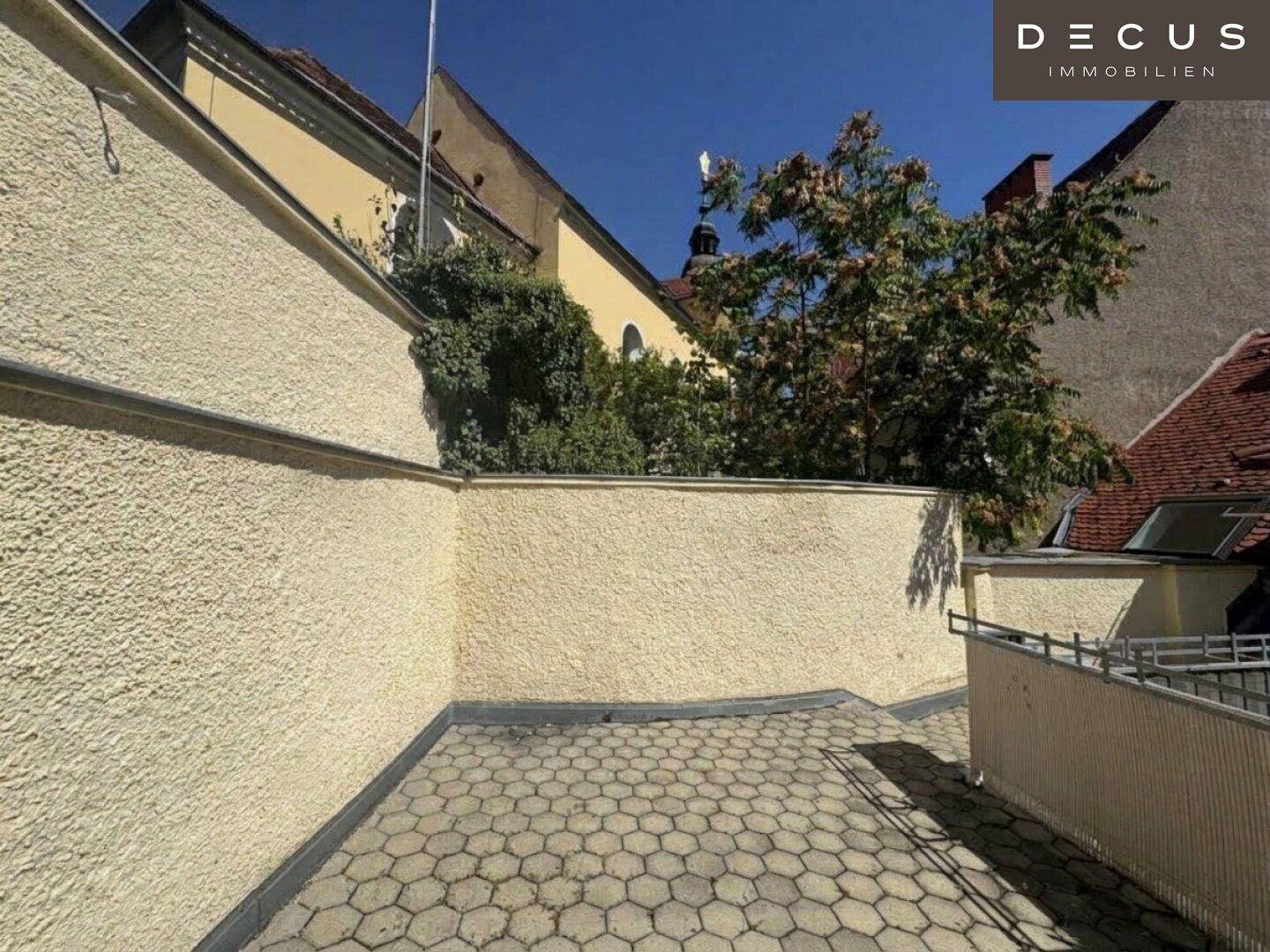 Gepflasterte Terrasse mit hohen Mauern und Blick auf umliegende Gebäude, sonnig und offen.