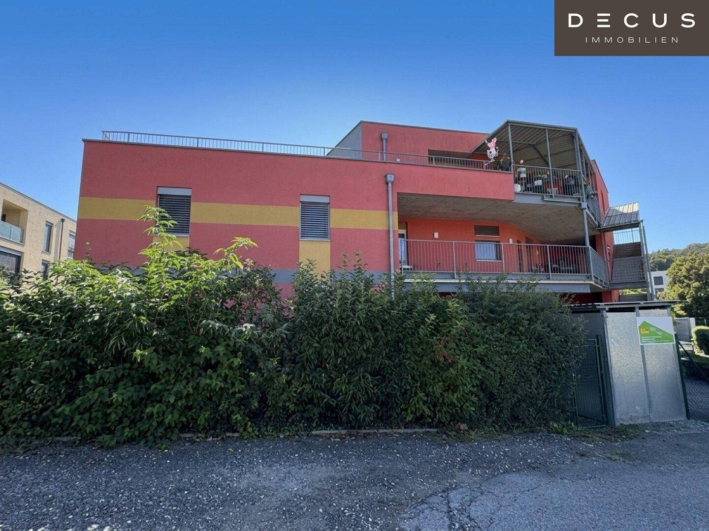 Seitenansicht des modernen Gebäudes mit roter Fassade, Balkonen und üppiger Bepflanzung im Vordergrund.