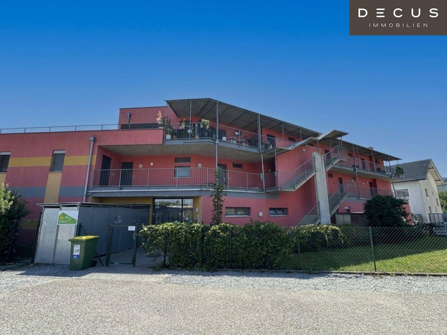 Weitläufige Ansicht des modernen Wohngebäudes mit roter Fassade und Balkonen unter blauem Himmel.