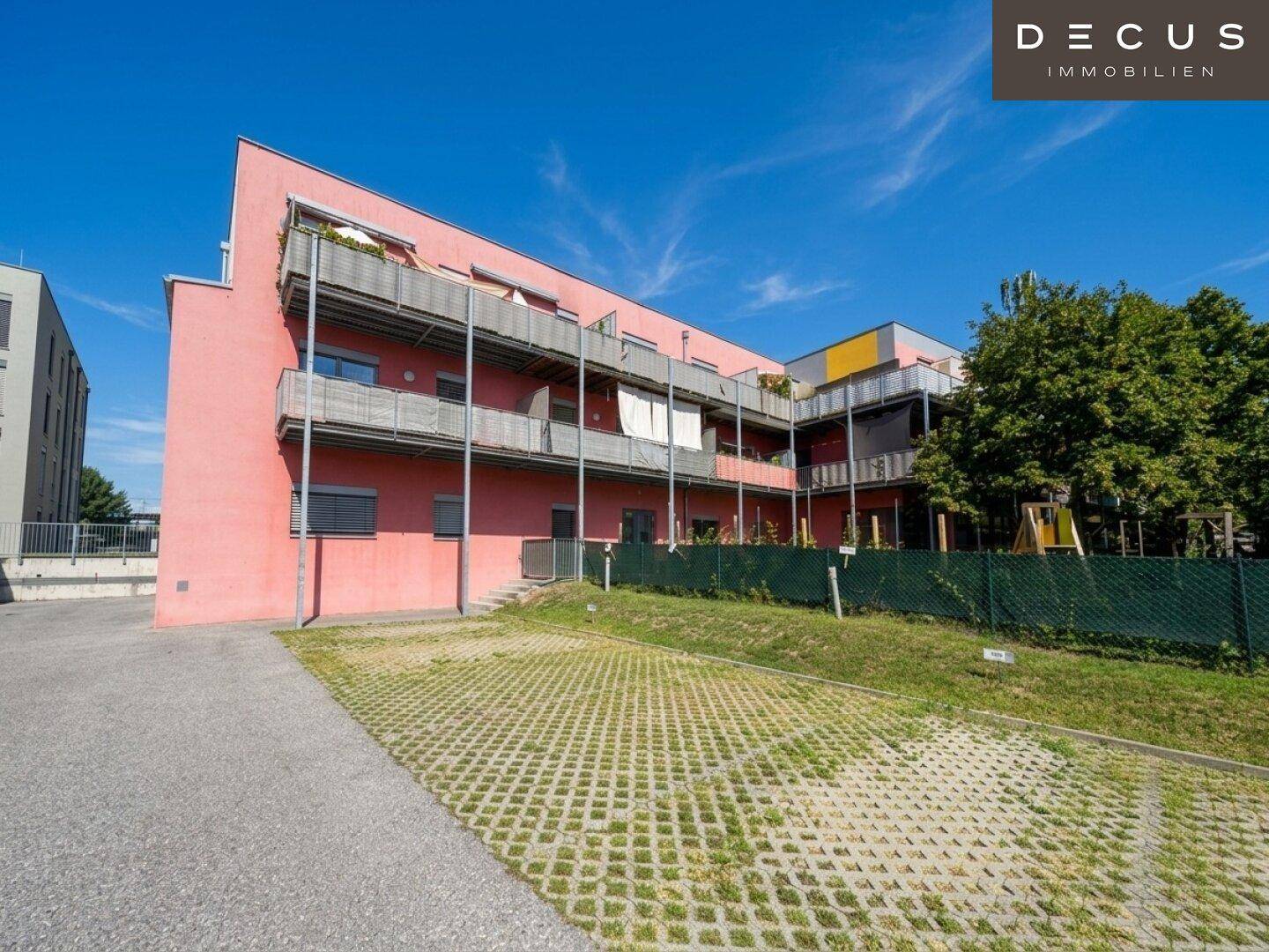 Modernes Wohngebäude mit roter Fassade und Balkonen, gesehen von einem Parkplatz mit Rasengittersteinen.