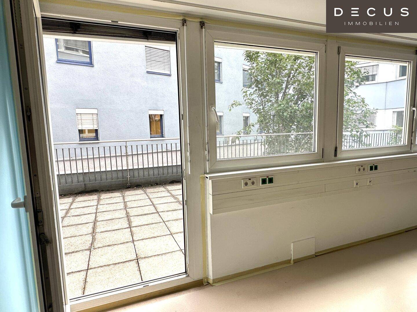Heller Innenraum mit großen Fenstern und Zugang zu einer weitläufigen, gepflasterten Terrasse.