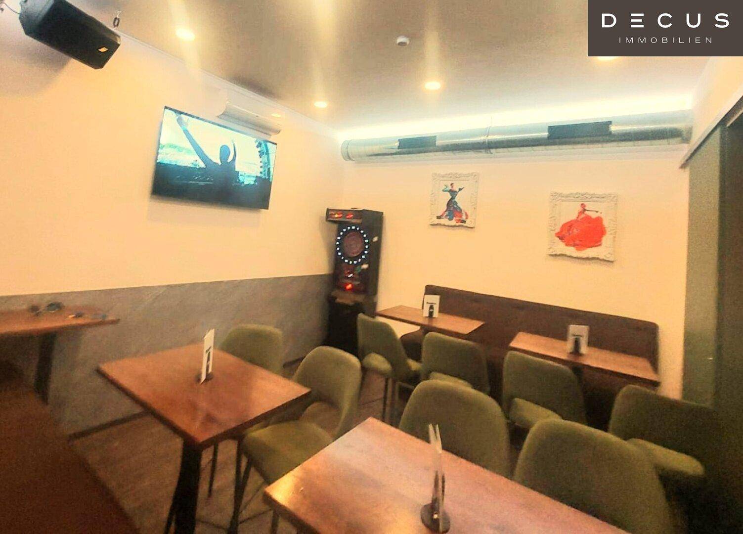 Unterhaltungsbereich mit Tischen, Sitzgelegenheiten, einem Dartautomaten und einem an der Wand montierten Fernseher.