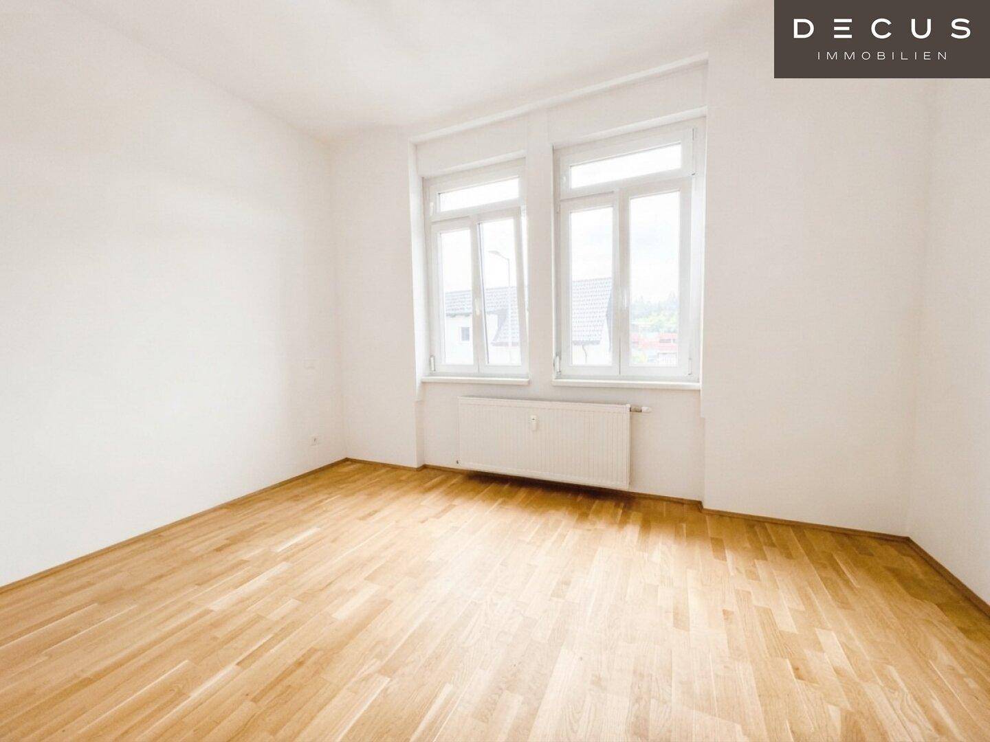 Helles Zimmer mit Holzboden und zwei großen Fenstern, bereit zur individuellen Gestaltung.