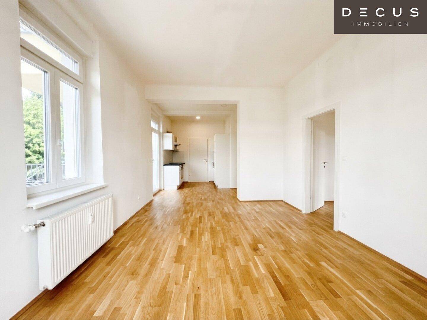Heller Wohnbereich mit Holzboden, großen Fenstern und einer integrierten Küchenzeile im Hintergrund.
