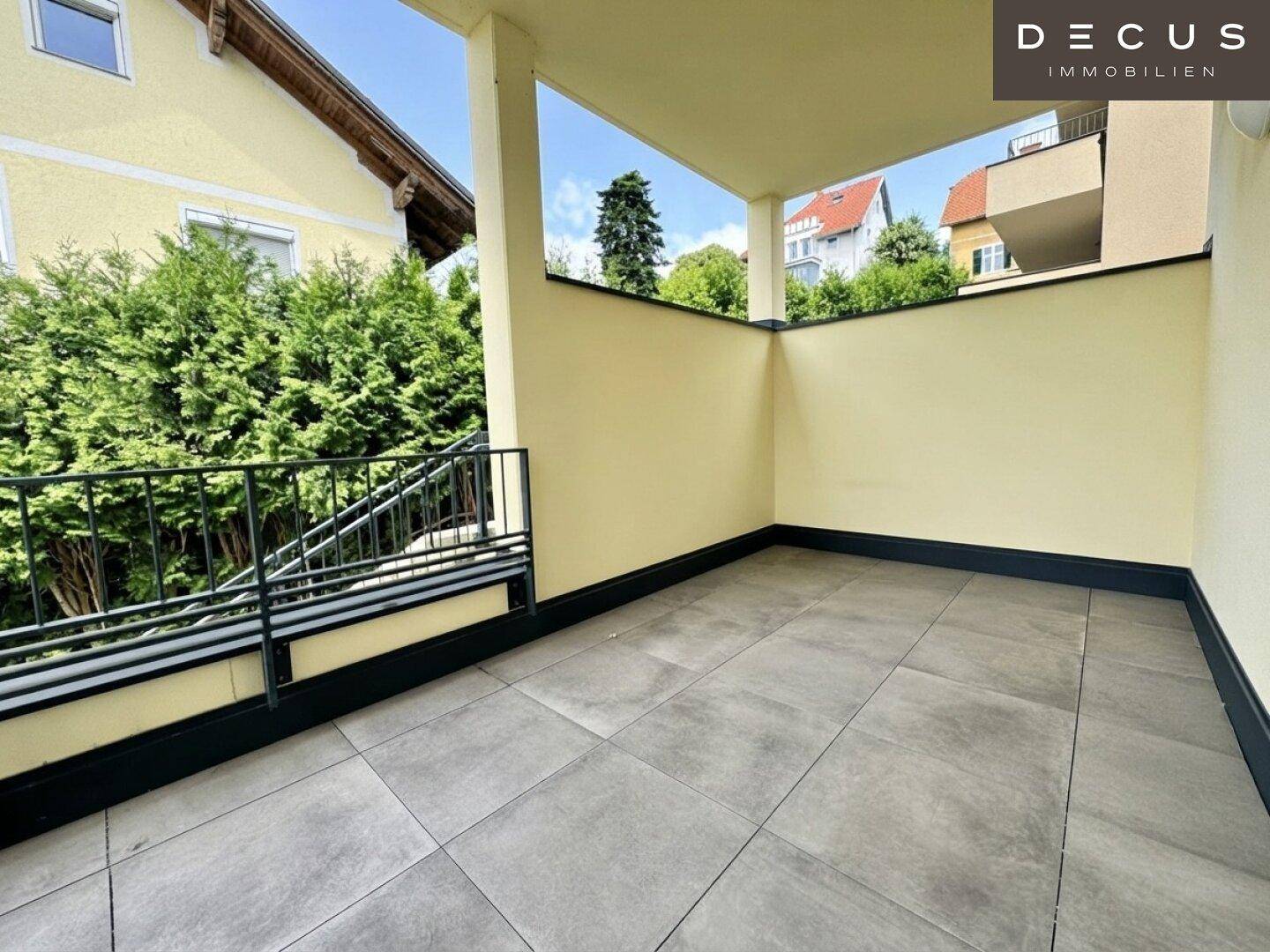 Geräumiger, überdachter Balkon mit modernen grauen Fliesen und Blick ins Grüne.