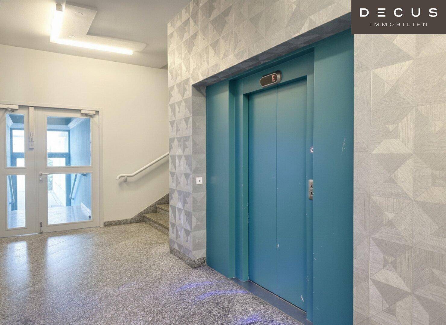 Heller Flur mit einem auffälligen blauen Aufzug und gemusterten Wänden, der zu den Treppen führt.