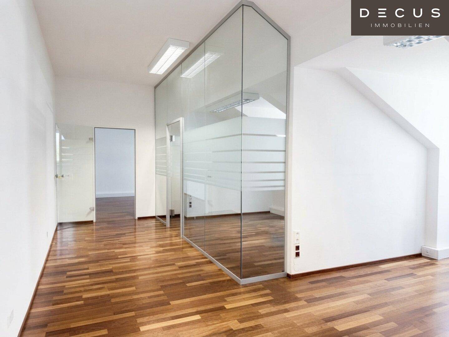 Modernes Büro mit Glasabtrennung, Holzboden und schrägen Wänden, das eine helle Arbeitsumgebung bietet.