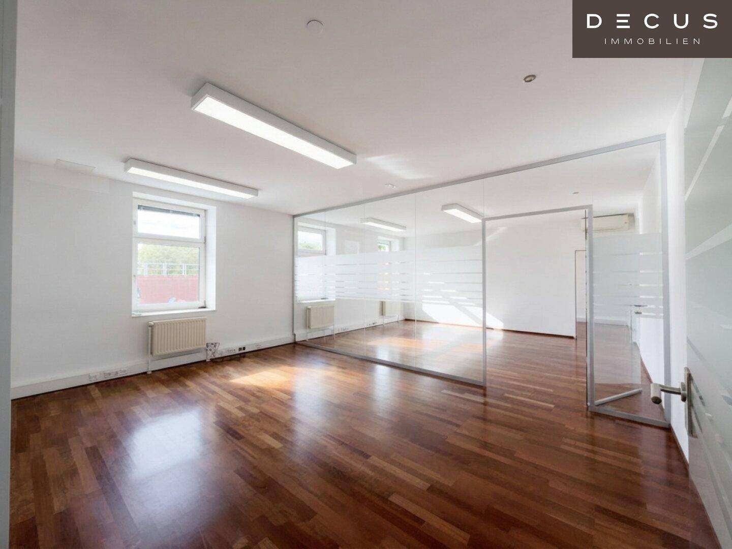 Großes, helles Büro mit Holzboden, Fenstern und einer Glaswand, die viel Tageslicht hereinlässt.