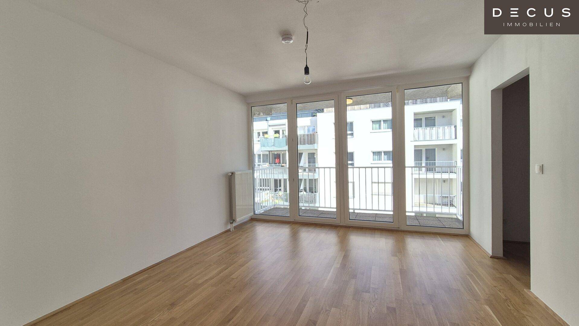 Geräumiger, unmöblierter Wohnraum mit Parkettboden und großen Fenstern zum Balkon.