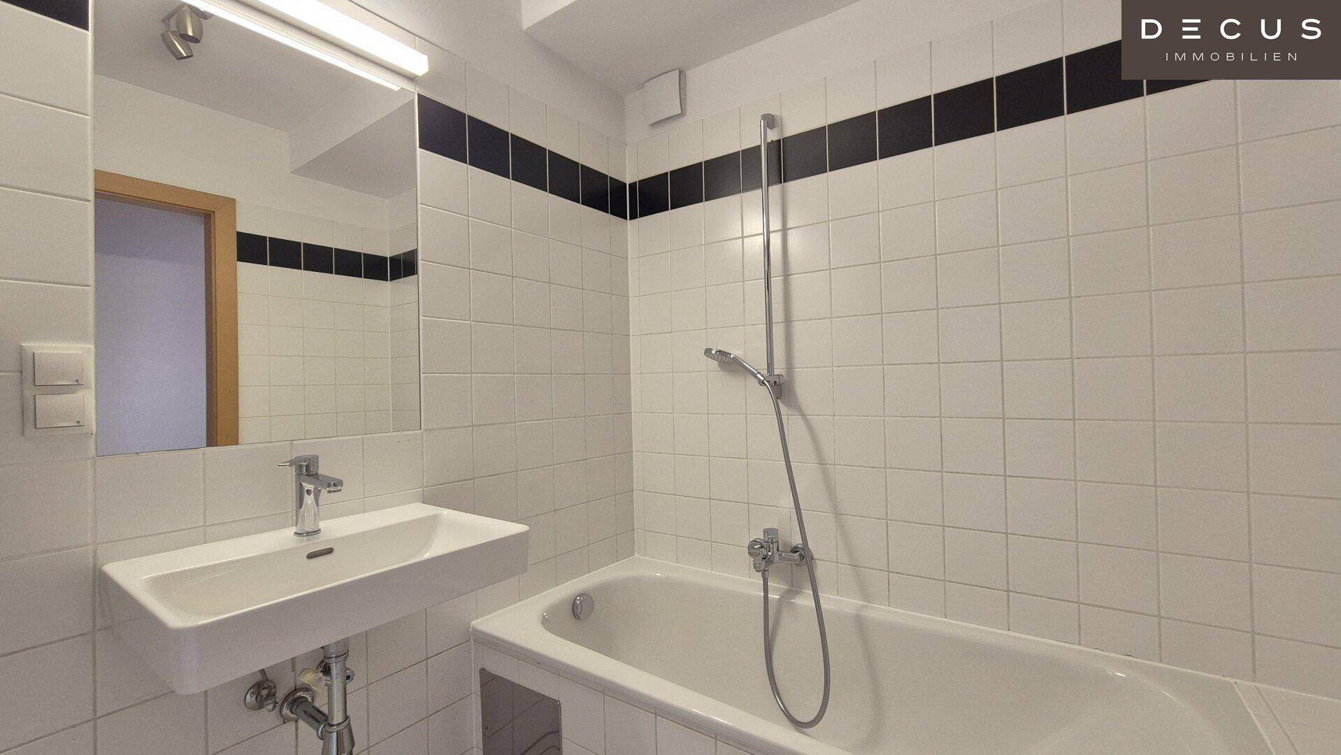 Badezimmer mit Badewanne, Waschbecken und weißen Fliesen, akzentuiert durch einen schwarzen Streifen.