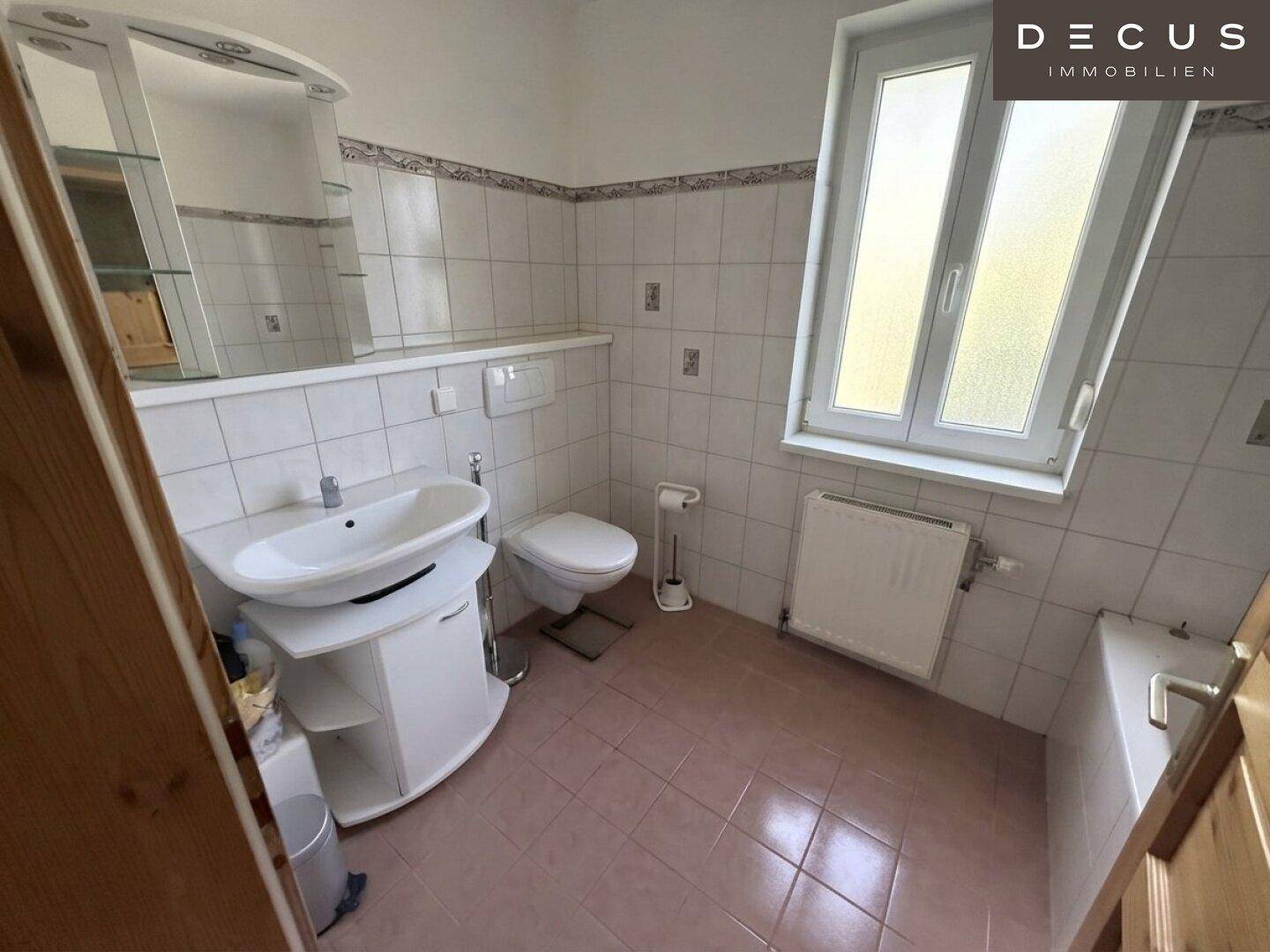 Badezimmer mit weißen Fliesen, Einzelwaschbecken, Toilette und Fenster, in gutem Zustand.