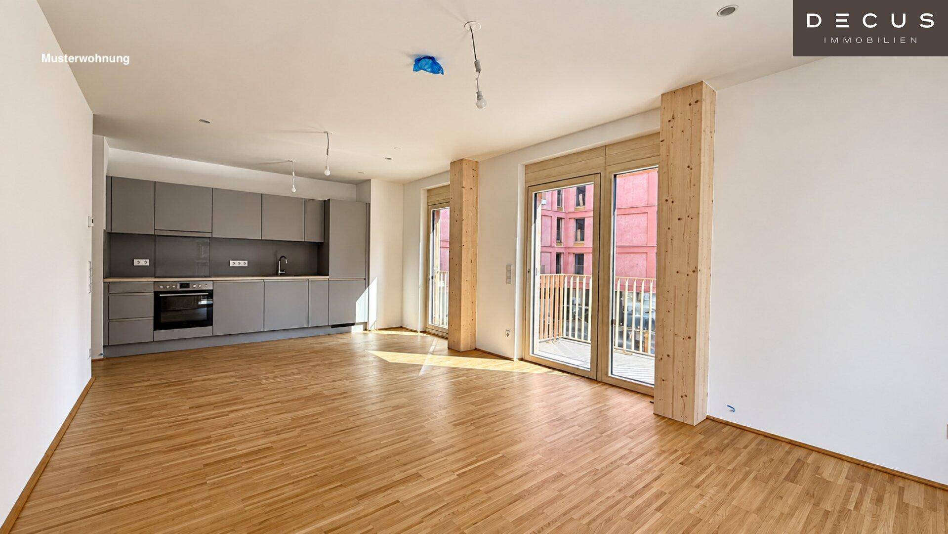Offener Wohnbereich einer Musterwohnung mit integrierter grauer Küche und Balkonzugang.