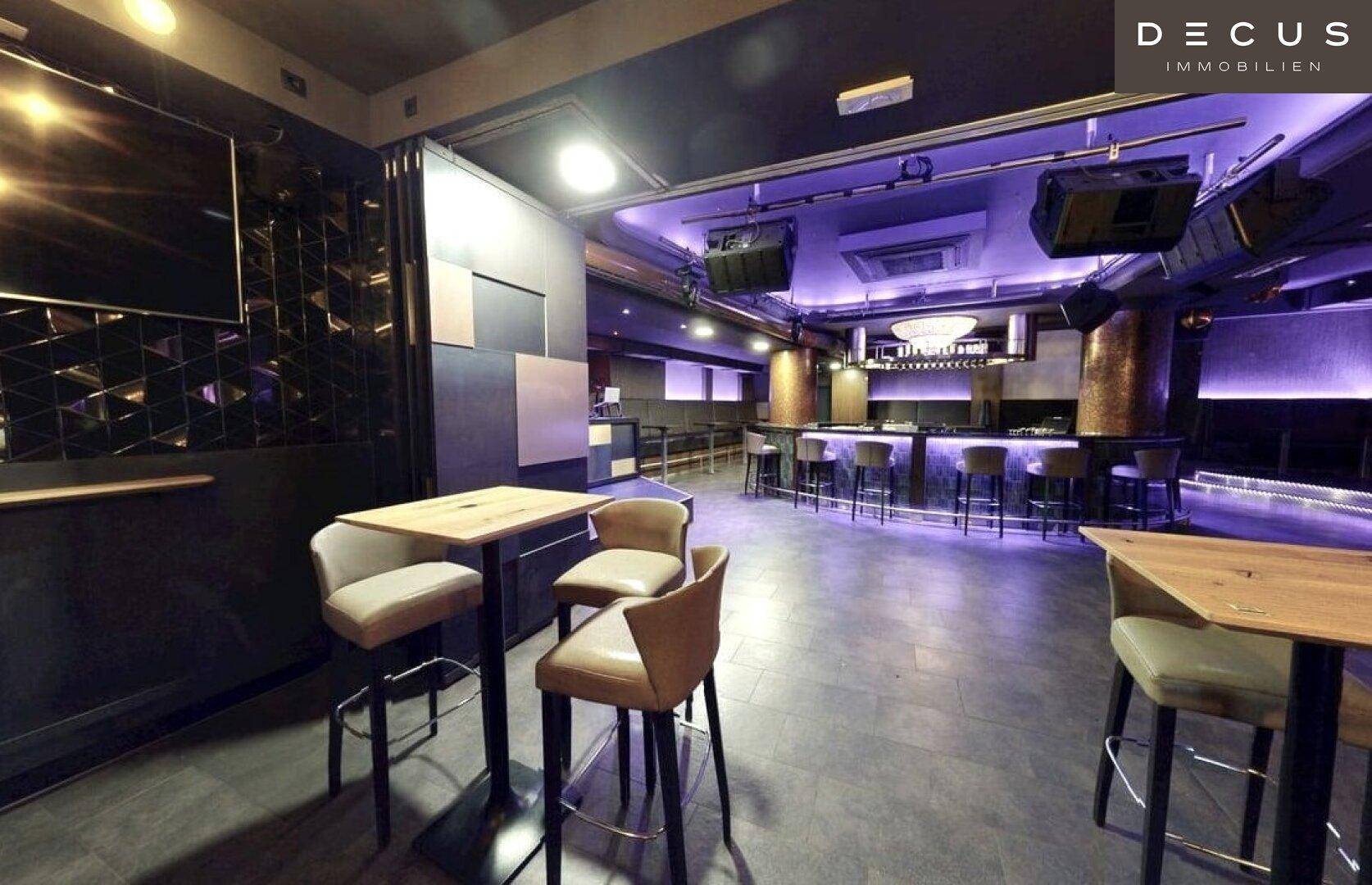 Weitläufiger Nachtclub mit dunklem Interieur, hohen Tischen und einer beleuchteten Bar im Hintergrund.