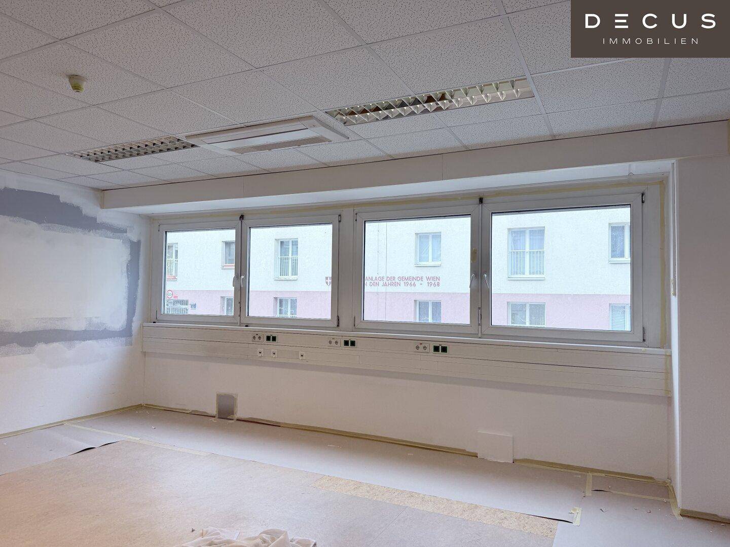Heller Büroraum mit großen Fenstern und Blick auf ein gegenüberliegendes Gebäude, Wände teilweise vorbereitet.