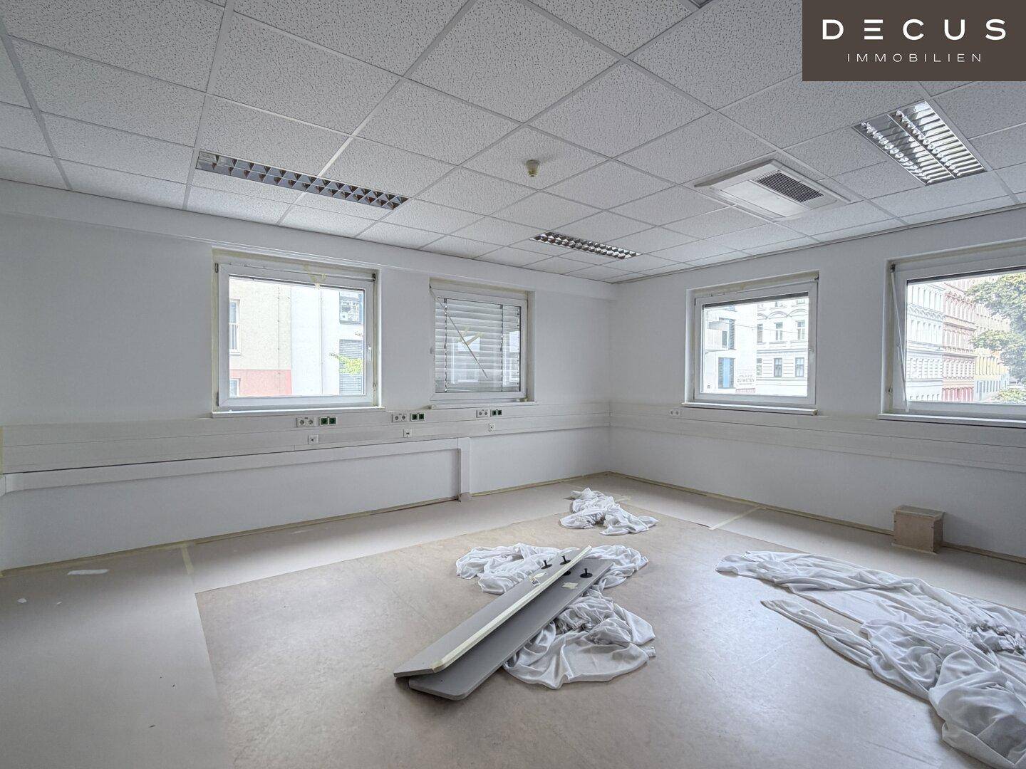 Leerer Büroraum mit mehreren Fenstern, Rasterdecke und einigen Bauabfällen auf dem Boden.