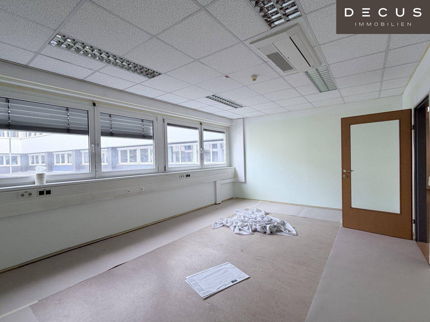 Geräumiger, leerer Büroraum mit großen Fenstern, Rasterdecke und einer offenen Holztür.