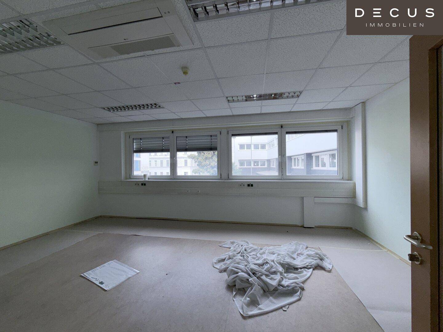 Leerer Büroraum mit Fenstern, Rasterdecke und einigen Materialien auf dem Boden, bereit zur Renovierung.