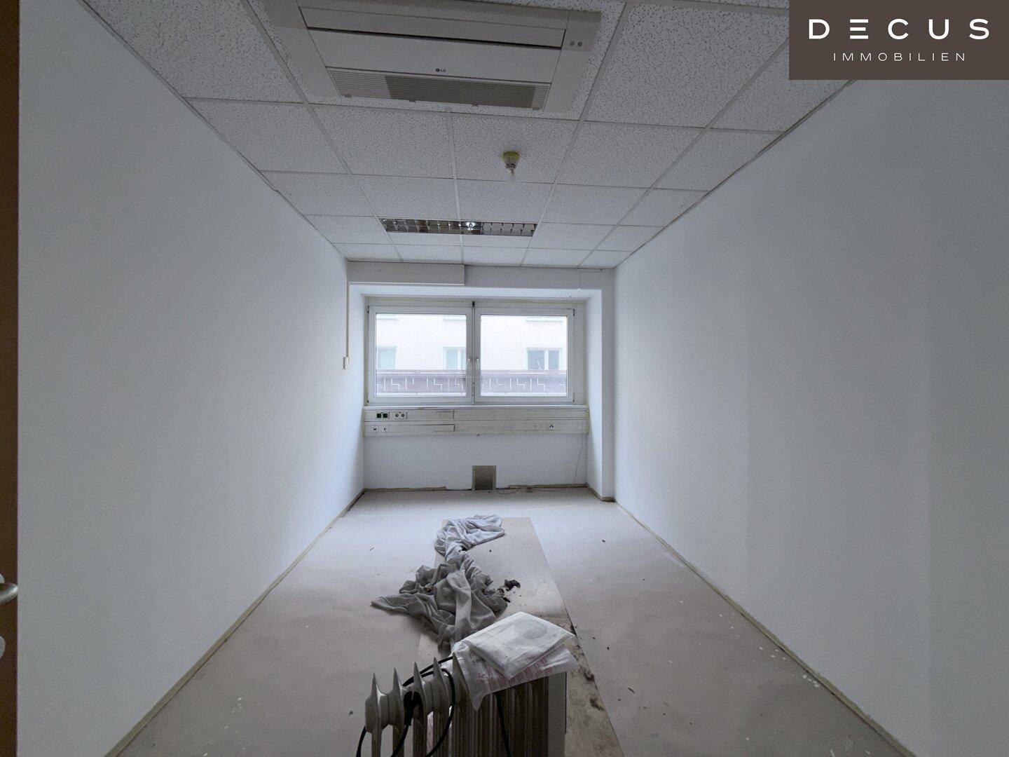Schmaler, leerer Büroraum mit einem Fenster, Rasterdecke und Heizkörper am Boden.