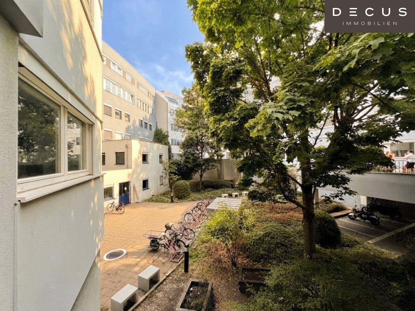Grüner Innenhof mit Bäumen, Fahrradstellplätzen und umliegenden Gebäuden unter blauem Himmel.