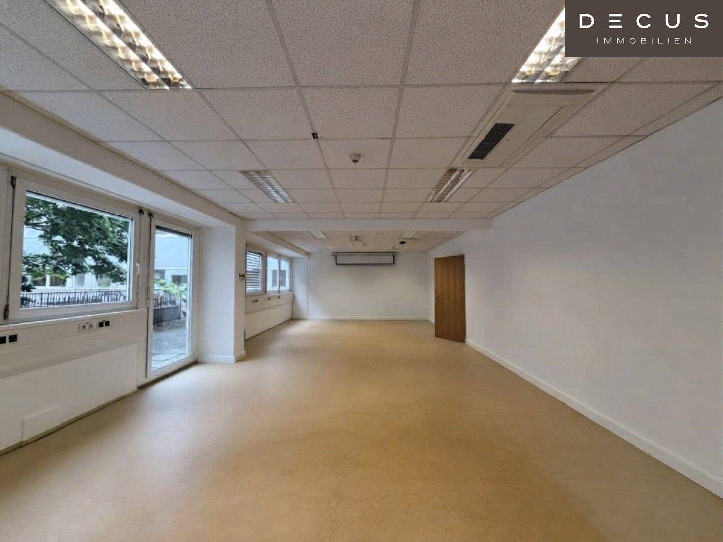 Langer, leerer Büroraum mit vielen Fenstern, die Tageslicht hereinlassen, und einer Rasterdecke.
