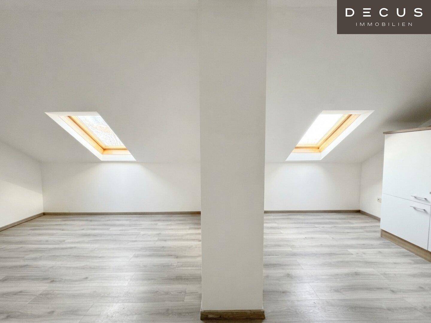Leerer Dachgeschossraum mit hellen Wänden, Laminatboden und zwei großen Dachfenstern für viel Tageslicht.