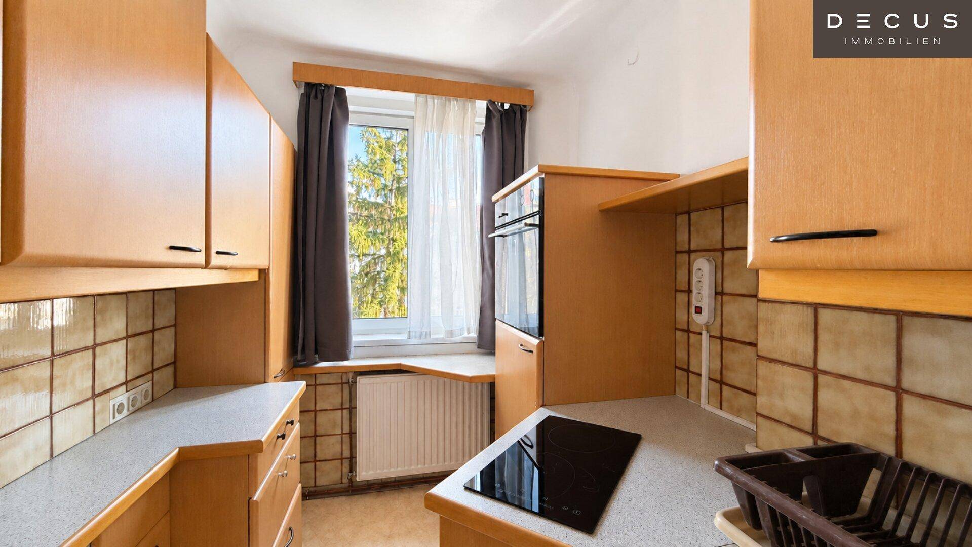 Funktionale Küche mit Holzschränken, Fliesenrückwand und Induktionskochfeld am Fenster, bietet viel Arbeitsfläche.