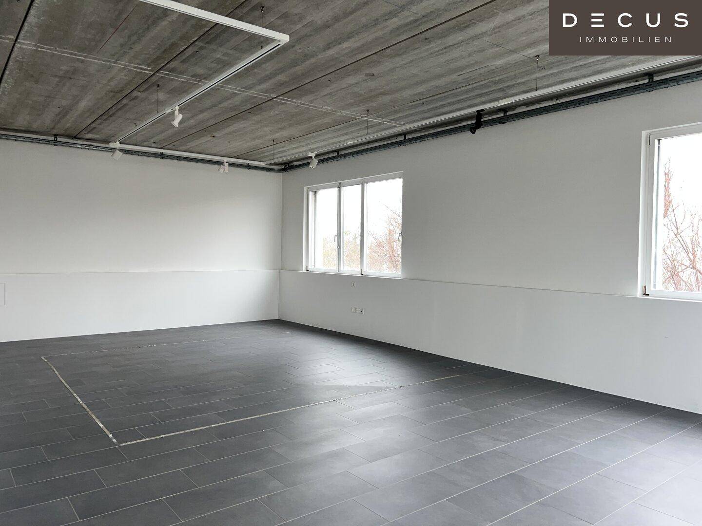 Großer, leerer Büroraum mit grauen Fliesen, weißen Wänden und einer sichtbaren Betondecke.