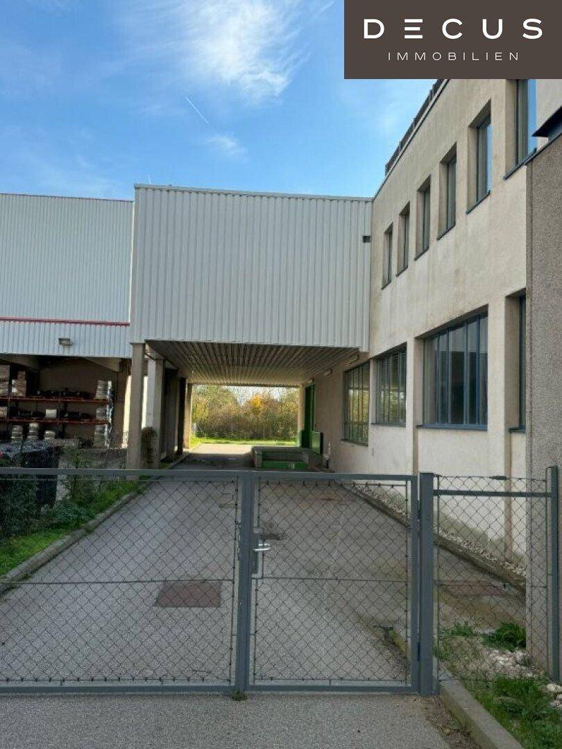 Zufahrt zum Gebäude mit Metallzaun und überdachtem Bereich, umgeben von Bäumen.