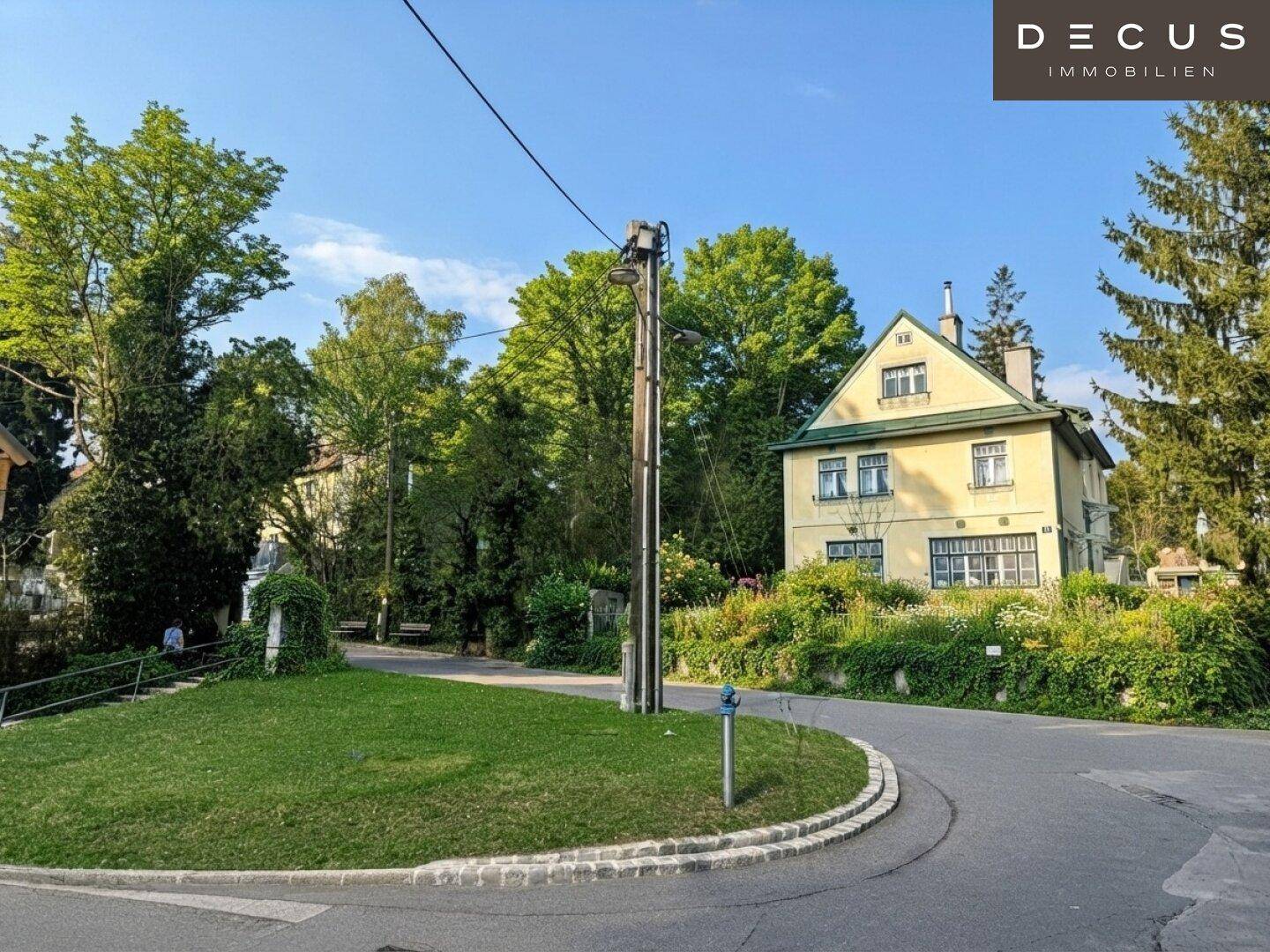 Straßenecke mit einer kleinen Grünfläche und einem traditionellen Haus im Hintergrund, umgeben von Bäumen.