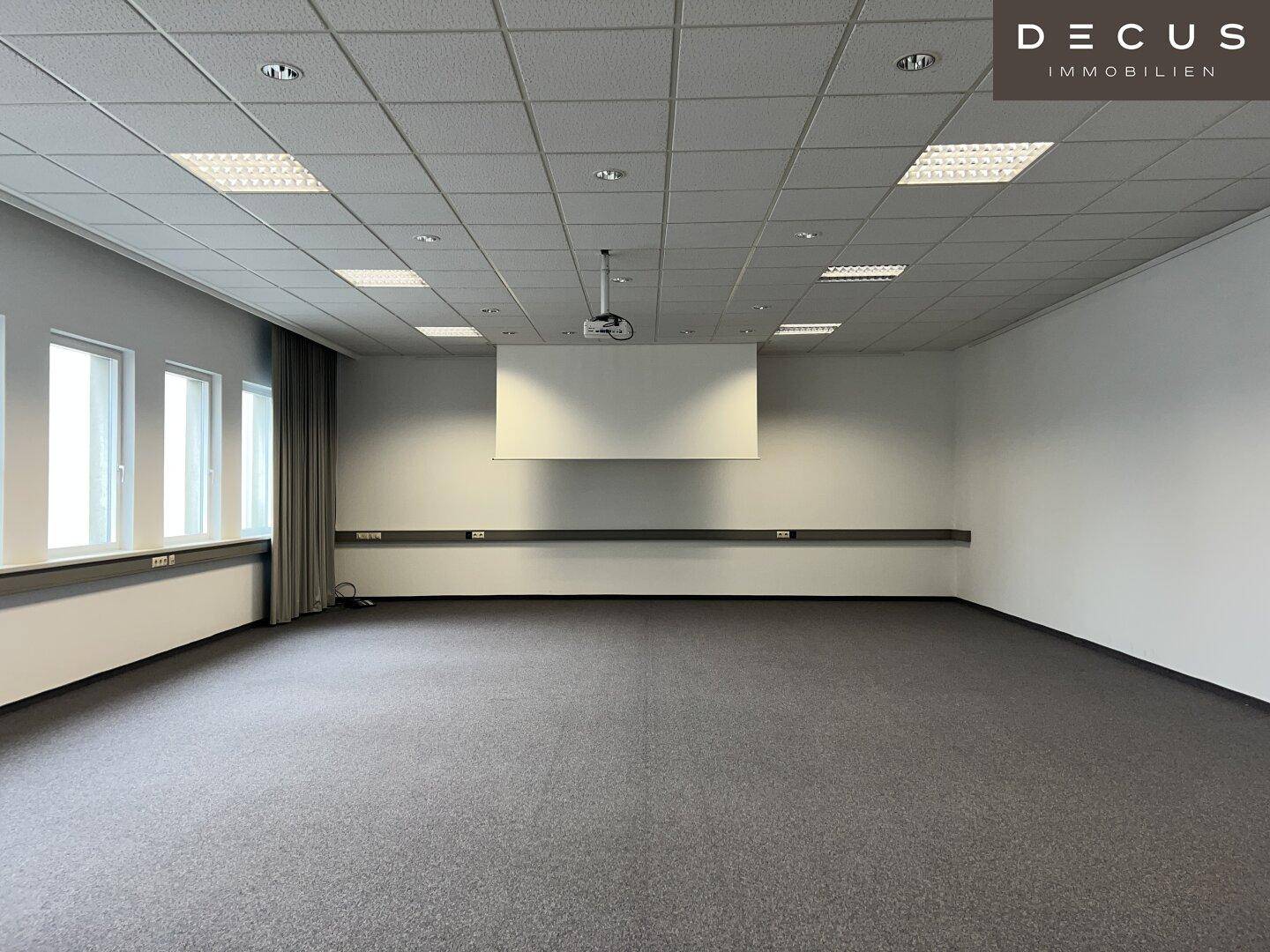 Großer, leerer Konferenzraum mit Teppichboden, abgehängter Decke und Projektorinstallation.