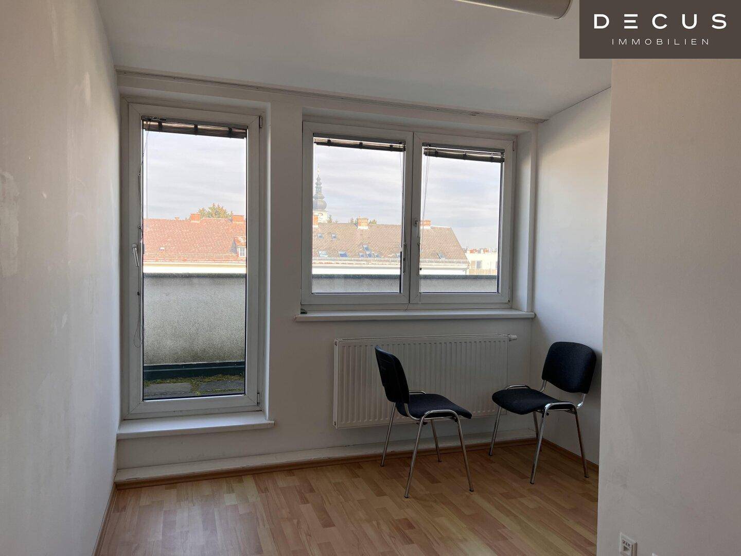 Heller Raum mit Laminatboden und großen Fenstern, die einen Blick auf die Dächer der Stadt bieten.