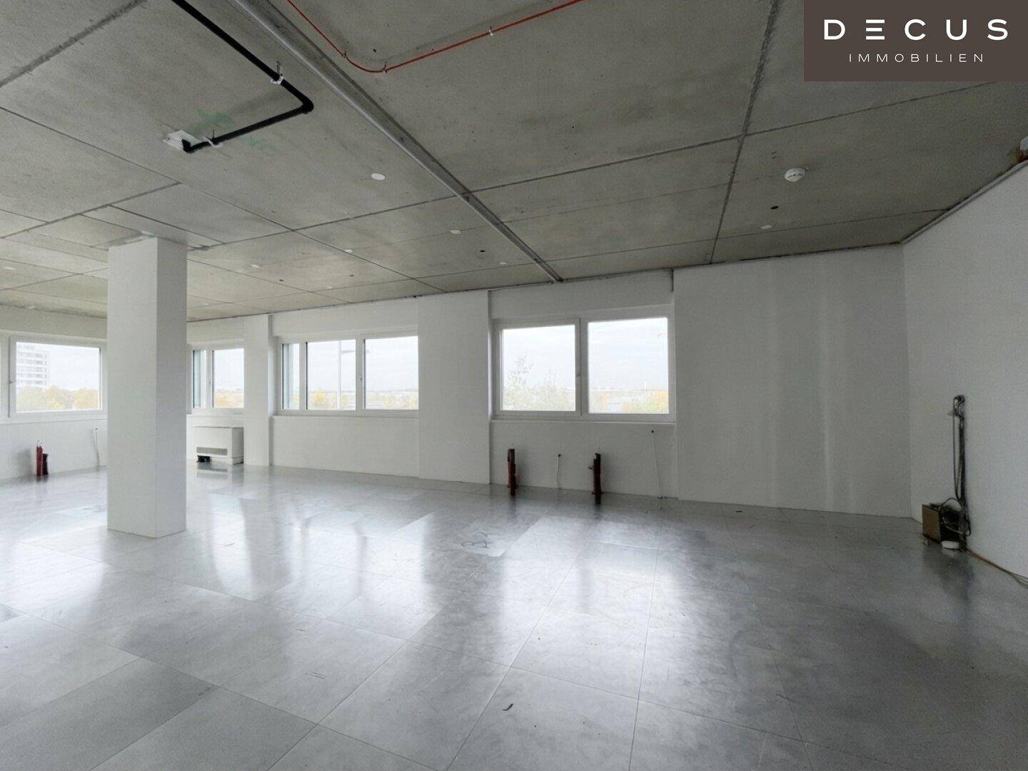 Großer, offener Büroraum mit vielen Fenstern, Betonboden und sichtbaren Deckeninstallationen.