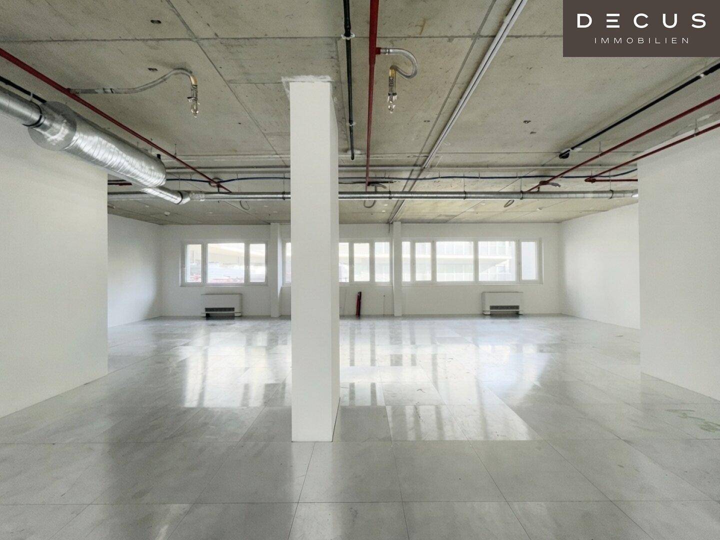Offener Bürobereich mit Säulen, vielen Fenstern und sichtbaren Lüftungsrohren an der hohen Decke.