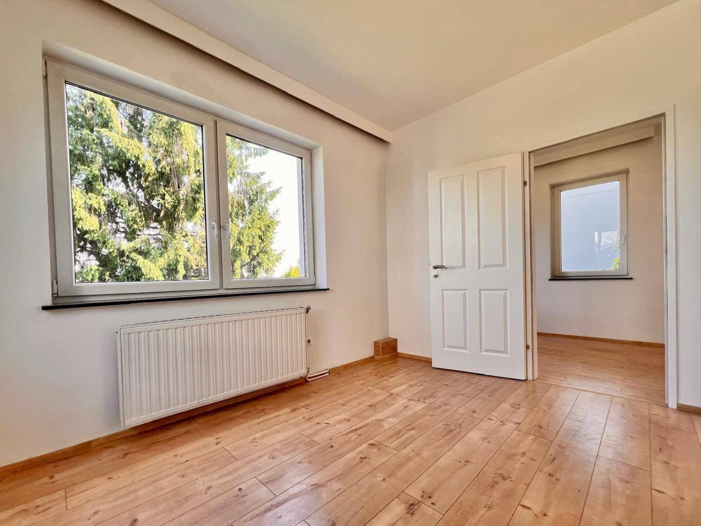 Geräumiges Zimmer mit Holzboden und großen Fenstern, die viel Tageslicht hereinlassen und Blick ins Grüne bieten.