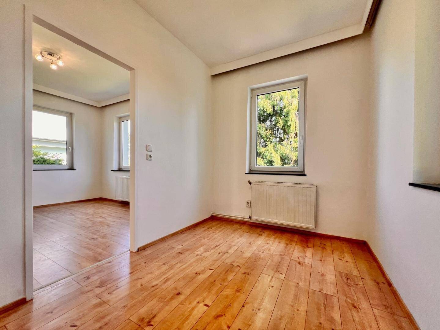 Lichtdurchflutetes Zimmer mit Holzboden und Blick ins Grüne durch die Fenster, bereit zur individuellen Gestaltung.