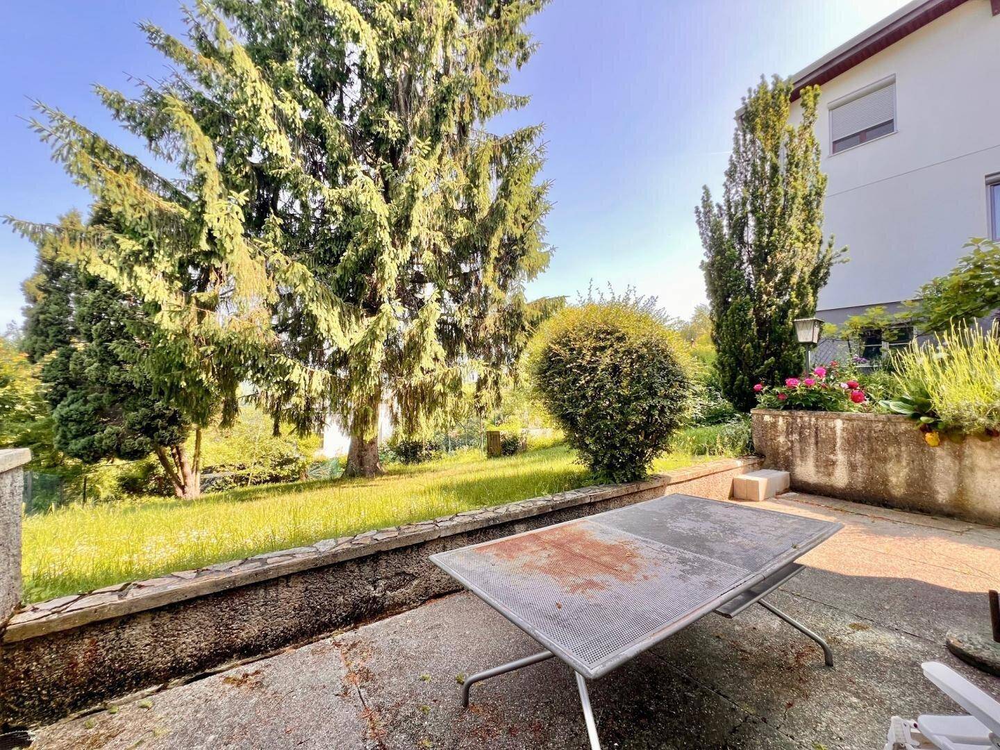 Terrasse mit einem Tisch und Blick auf den Garten mit altem Baumbestand, ideal für Mahlzeiten im Freien.
