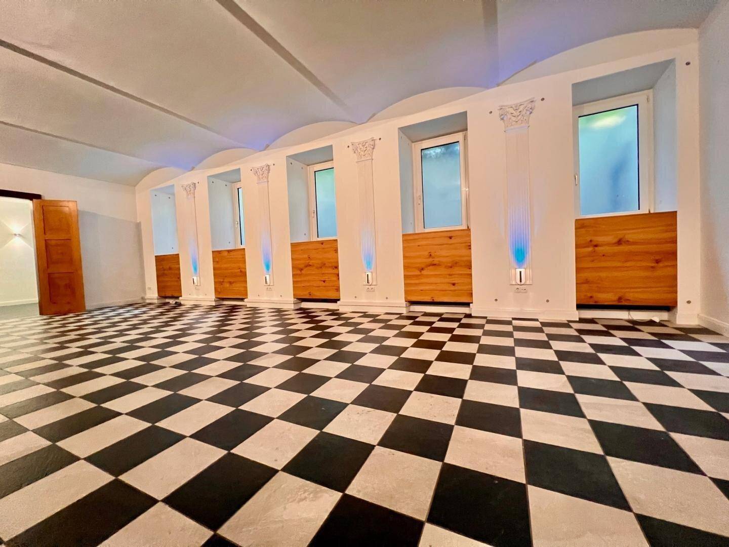 Geräumiger Raum mit schwarz-weißem Fliesenboden, gewölbter Decke und mehreren Fenstern mit dekorativen Säulen.